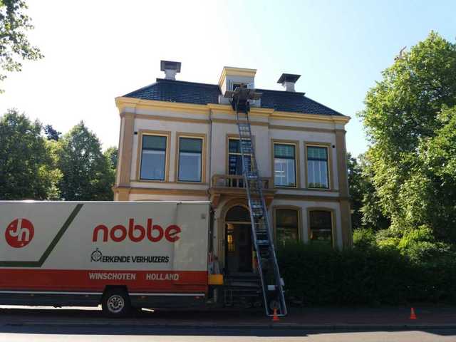 Voor een statig Herenhuis, omgeven met bomen en struiken staat een verhuiswagen van Nobbe. Onze verhuislift staat aan een raam op de tweede etage, waar de een deel van de spullen voor deze particuliere verhuizing moeten worden geplaatst.